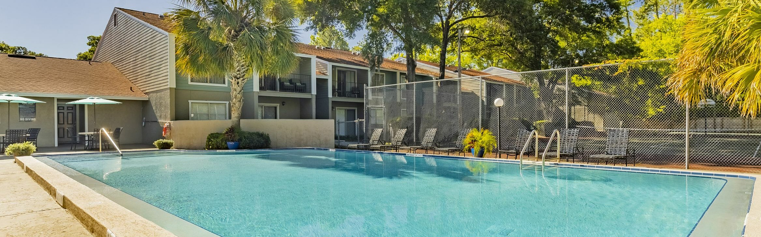 Inviting outdoor swimming pool area with sunlit lounging spaces, surrounded by lush greenery and modern apartment exteriors.