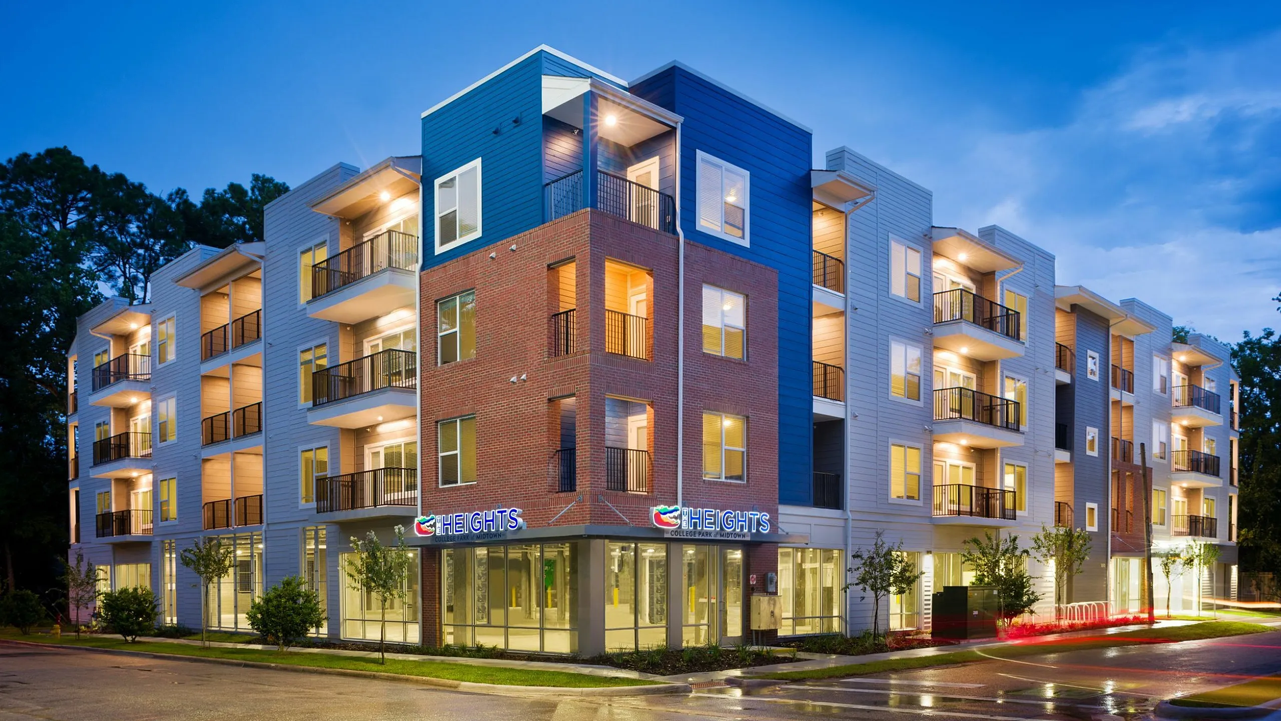 Modern multi-story apartment building with illuminated balconies at dusk, surrounded by lush greenery.