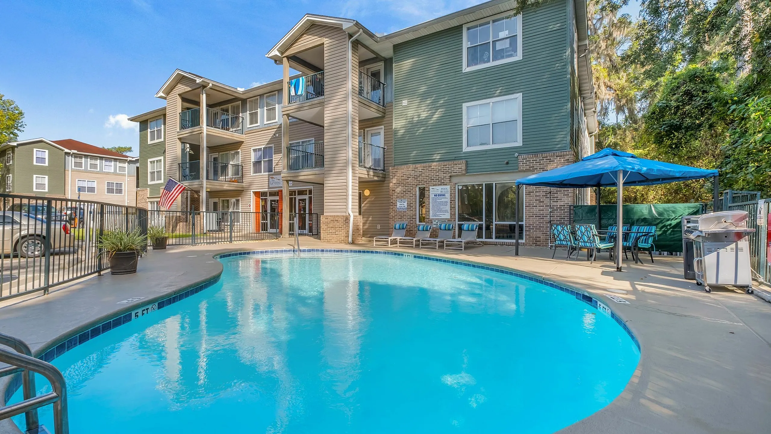 Bright apartment complex with refreshing pool, lounge chairs, and shaded seating area under blue skies.