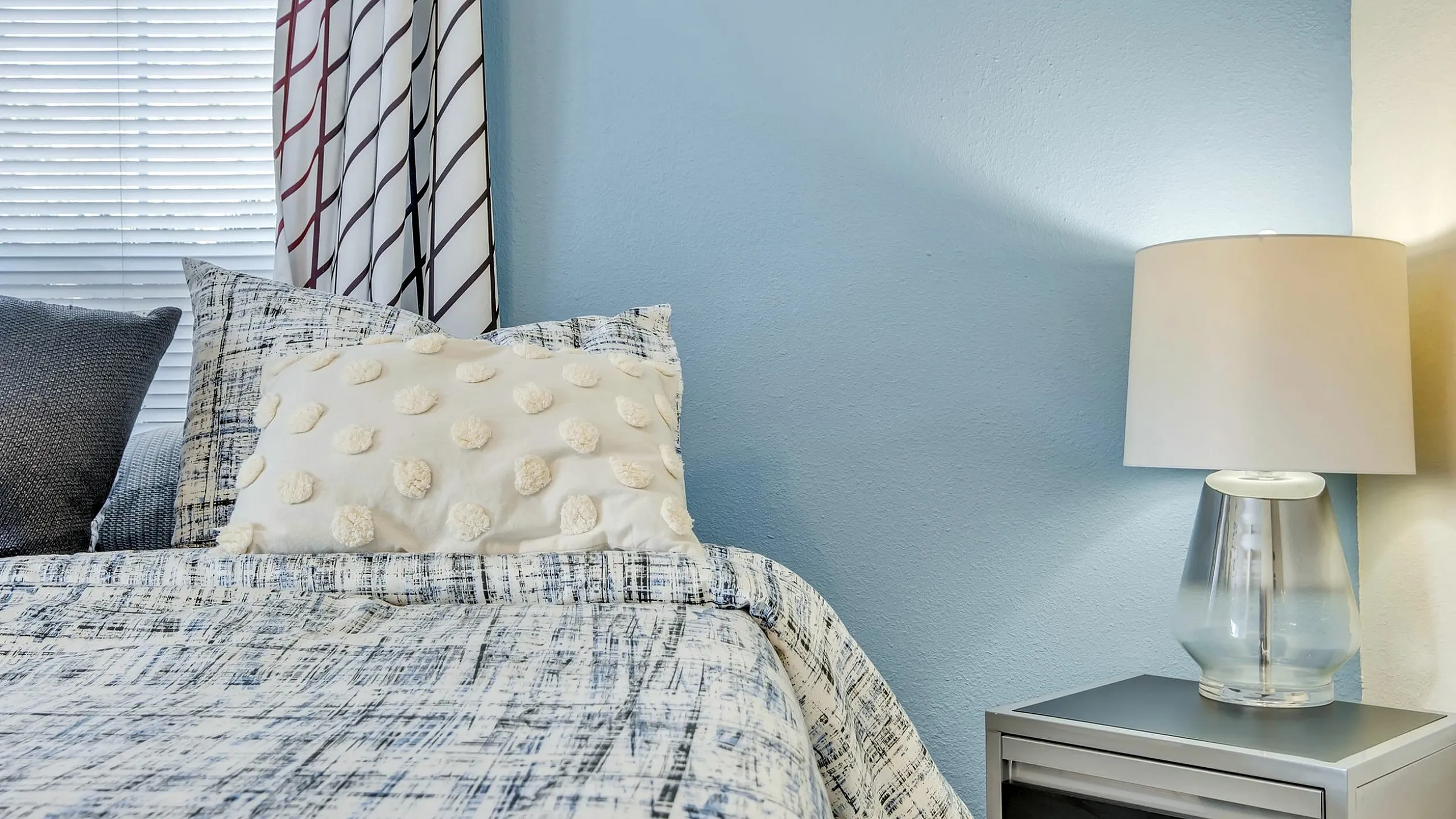 Cozy bedroom corner with elegant blue accent wall, chic patterned curtains, soft bedding, and ambient lamp on a modern nightstand.