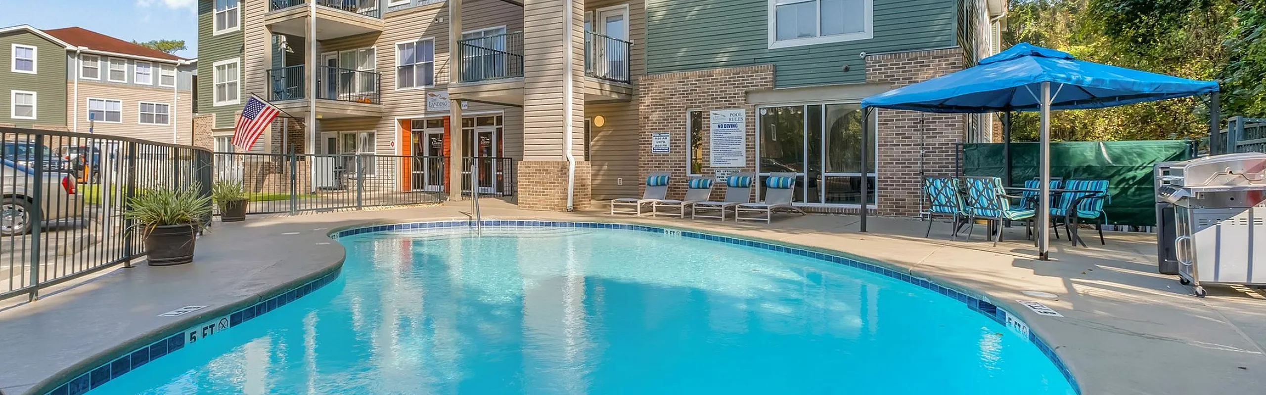 Inviting apartment complex with a sparkling outdoor pool, sun loungers, and shaded seating area under a clear blue sky.