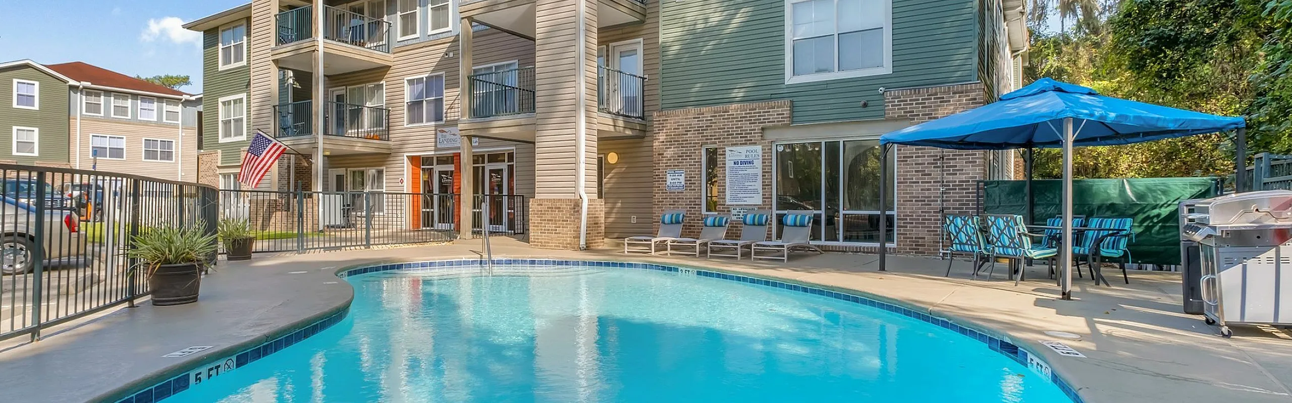 Sunlit apartment complex with balconies overlooking an inviting outdoor swimming pool, surrounded by lush greenery and a relaxing patio area with a barbecue grill.