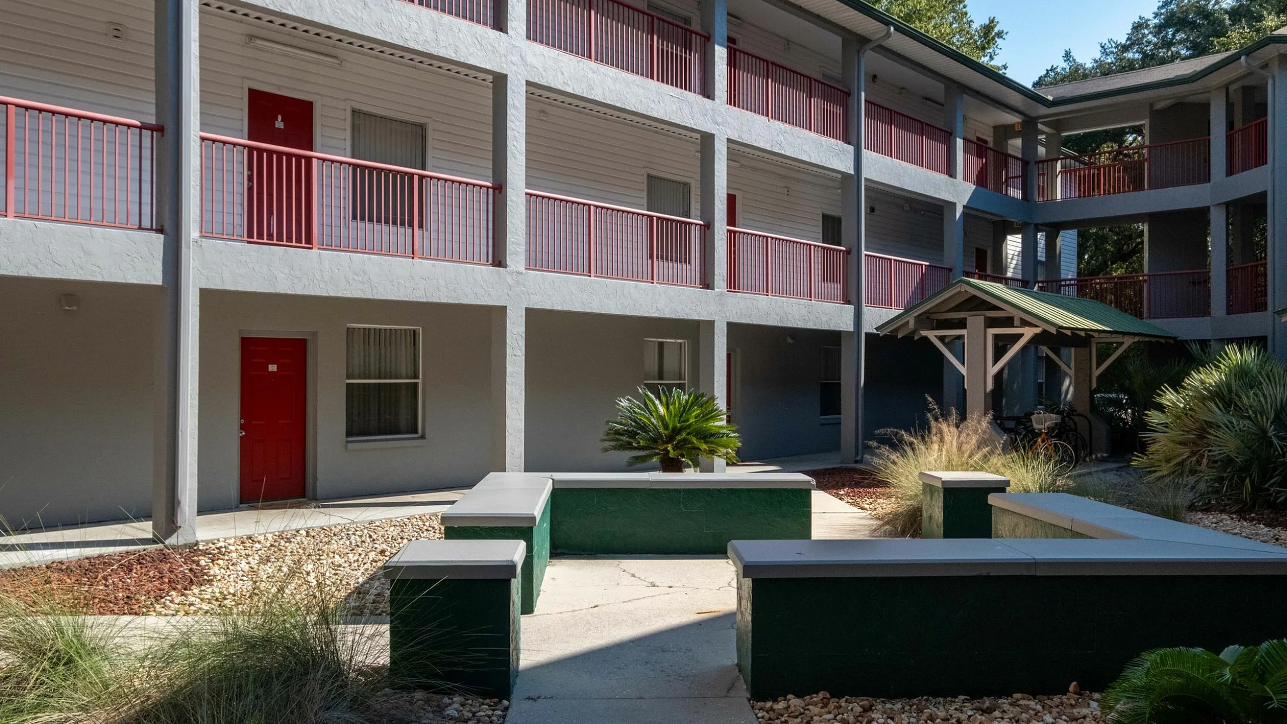 Charming apartment exterior featuring a modern three-story design with red accents, lush landscaping, and a relaxed courtyard setting.