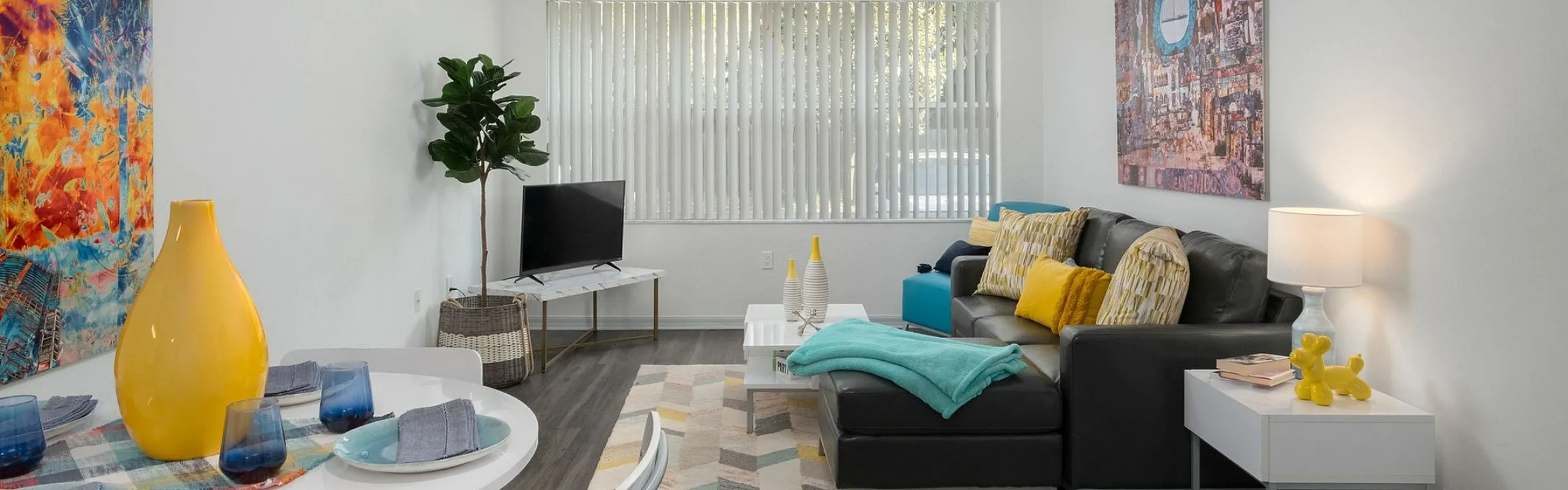 Vibrant living and dining area with modern decor, featuring colorful wall art, a cozy grey sofa adorned with yellow and teal accents, and a sleek white dining table set. Abundant natural light fills the room through large windows, highlight