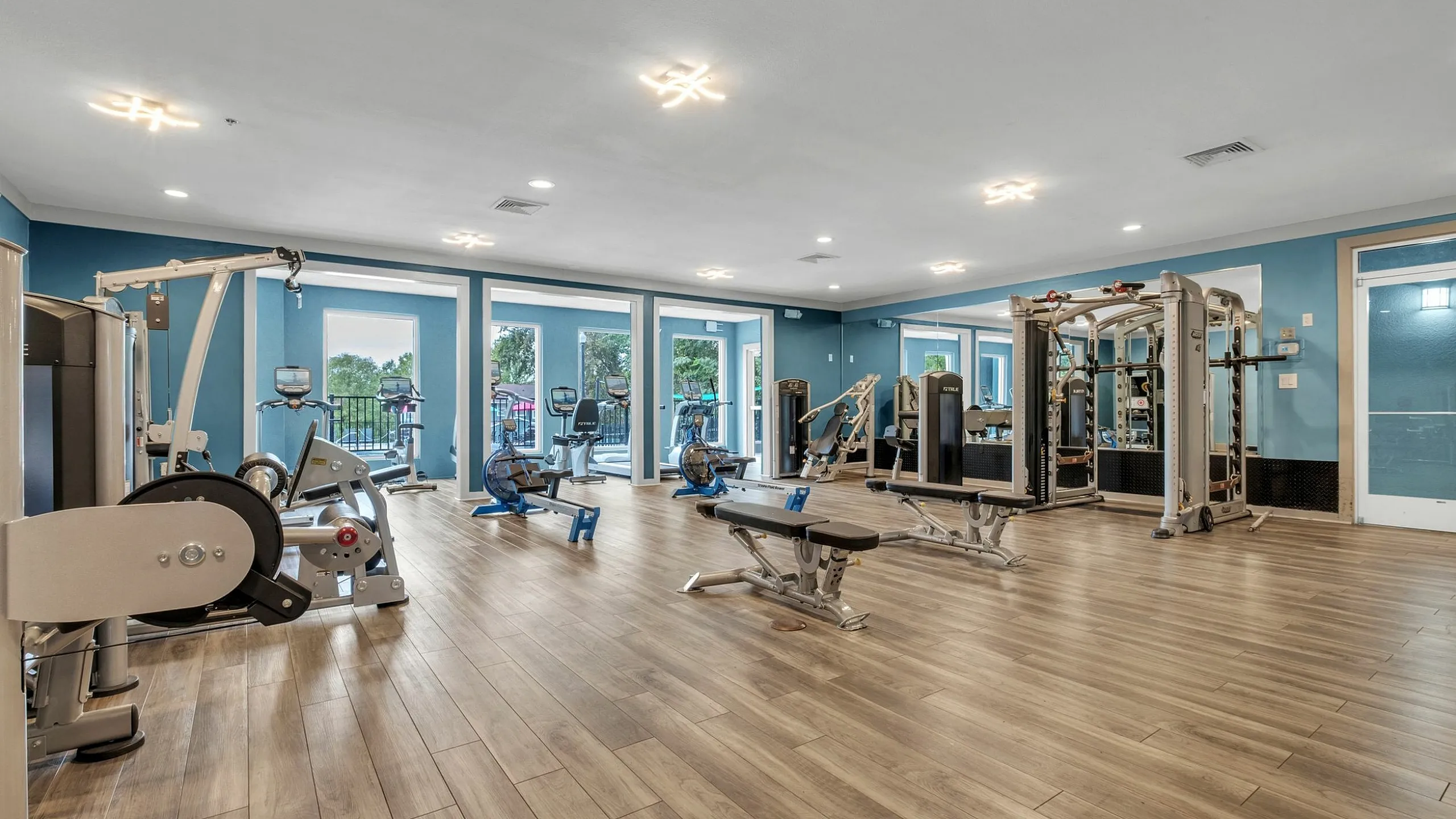 Expansive fitness center with state-of-the-art equipment, warm wooden flooring, and abundant natural light from large windows.