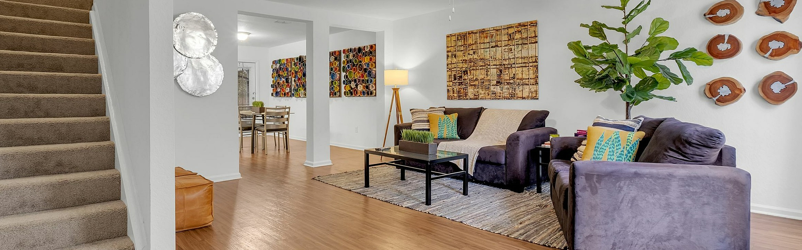 Inviting living room with cozy furniture, vibrant artwork, and modern decor, leading to a stylish dining area. Natural wood floors and lush greenery enhance the serene ambiance.