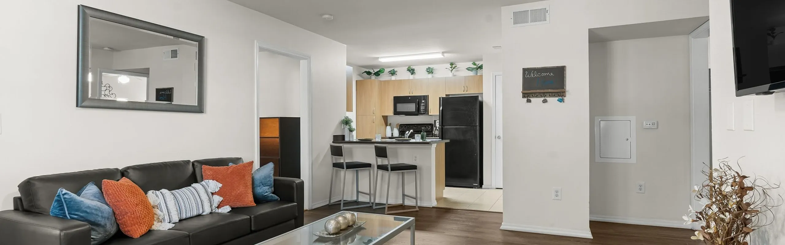 Contemporary open-plan living room with sleek black furniture, vibrant accent pillows, and a modern kitchen in the background.