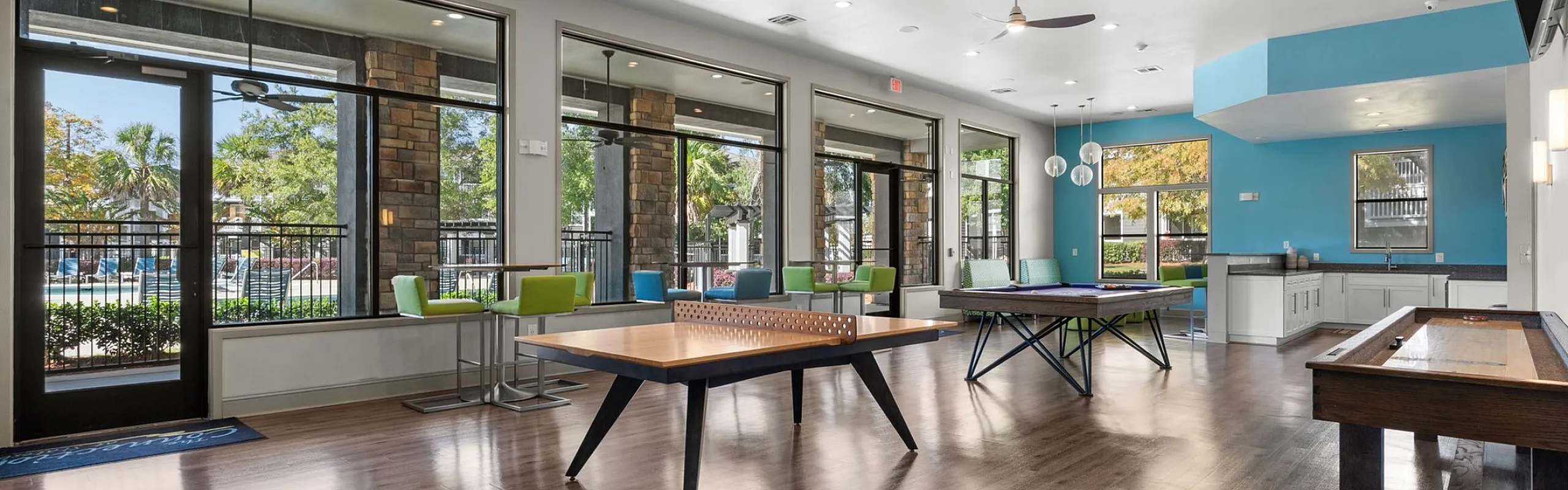Vibrant recreational room with stylish game tables, sleek modern ceiling fans, large windows, and a cozy kitchen area, perfect for social gatherings.