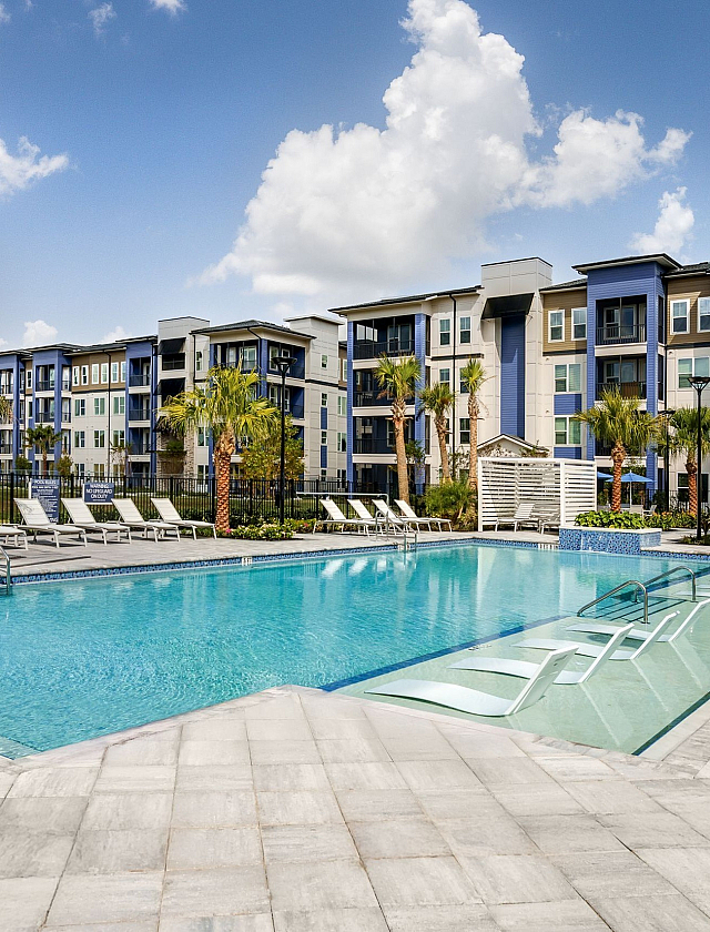 Luxurious apartment complex featuring a pristine swimming pool with modern lounge chairs, surrounded by lush greenery and elegant multi-story buildings under a vibrant blue sky.