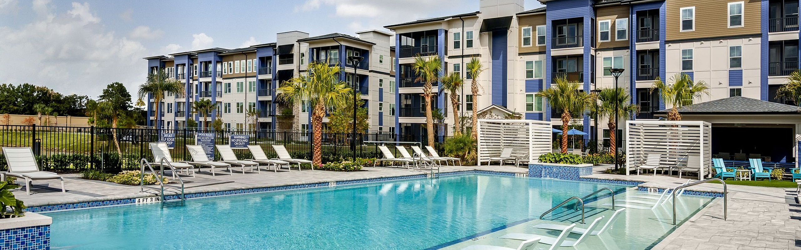 Luxurious apartment complex featuring a stunning pool with poolside lounge chairs, surrounded by modern architecture and lush palm trees.