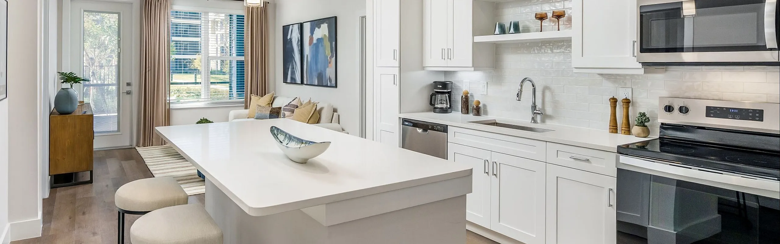Modern kitchen and living area with sleek white cabinetry, spacious island, and elegant d&eacute;cor, boasting bright natural light through large windows.
