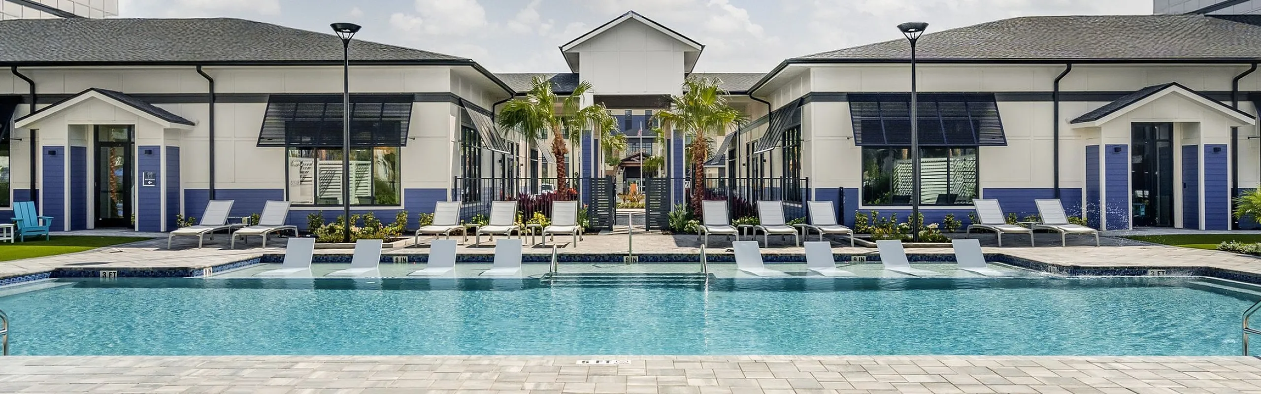 Luxurious apartment complex with a sparkling swimming pool and stylish lounge chairs, enclosed by contemporary buildings under a clear blue sky.