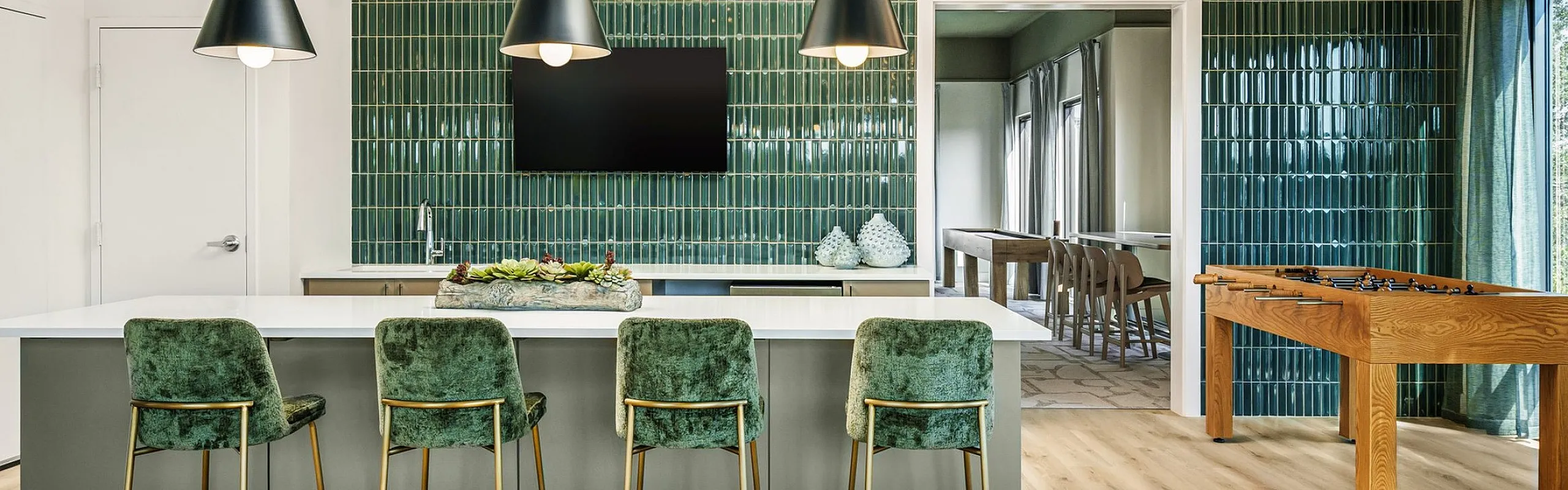 Modern kitchen and entertainment area with sleek green-tiled walls, stylish pendant lighting, and vibrant velvet bar stools, leading to a cozy game room with a foosball table.