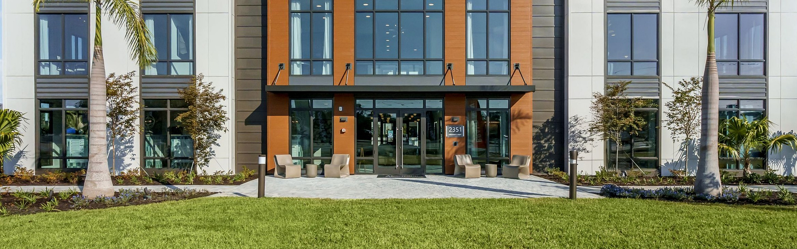 Modern apartment building exterior with sleek architectural design, large windows, and inviting entrance surrounded by lush greenery and palm trees.