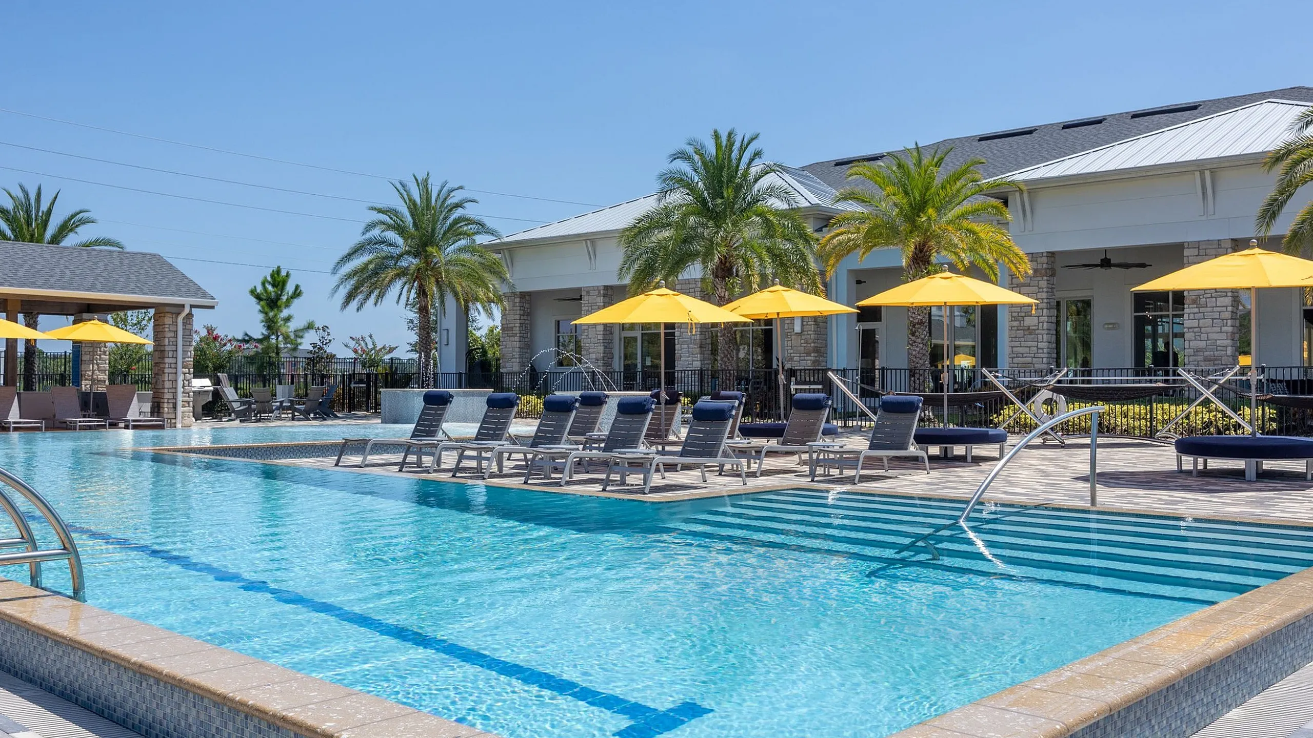 Luxurious apartment complex featuring a sparkling pool, sun loungers, and vibrant yellow umbrellas, surrounded by palm trees and elegant architecture.