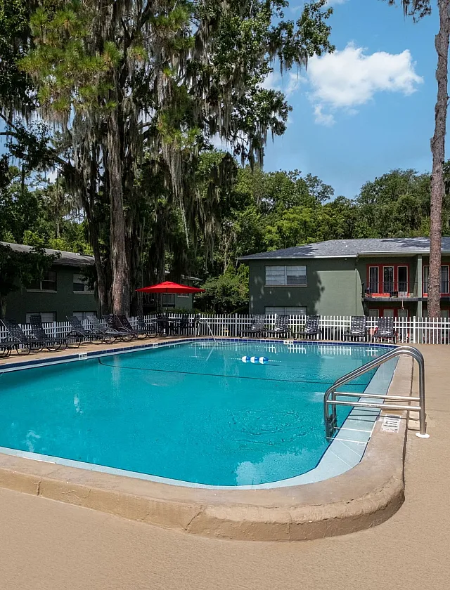 Inviting outdoor pool area surrounded by lush greenery and modern apartments with loungers and vibrant red umbrellas.
