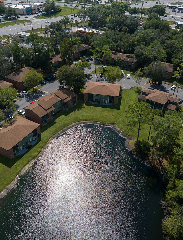 Charming aerial view of a serene apartment complex nestled by a shimmering lake, surrounded by lush greenery, with spacious units and inviting community pool.