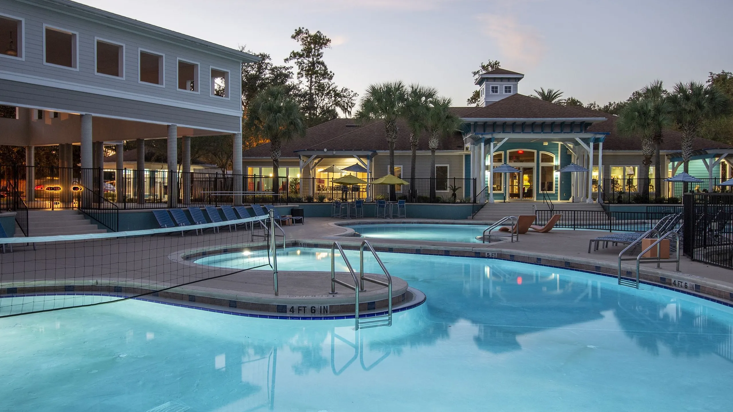 Inviting apartment complex with a beautifully lit swimming pool area, surrounded by stylish lounge chairs and modern architecture under a serene evening sky.