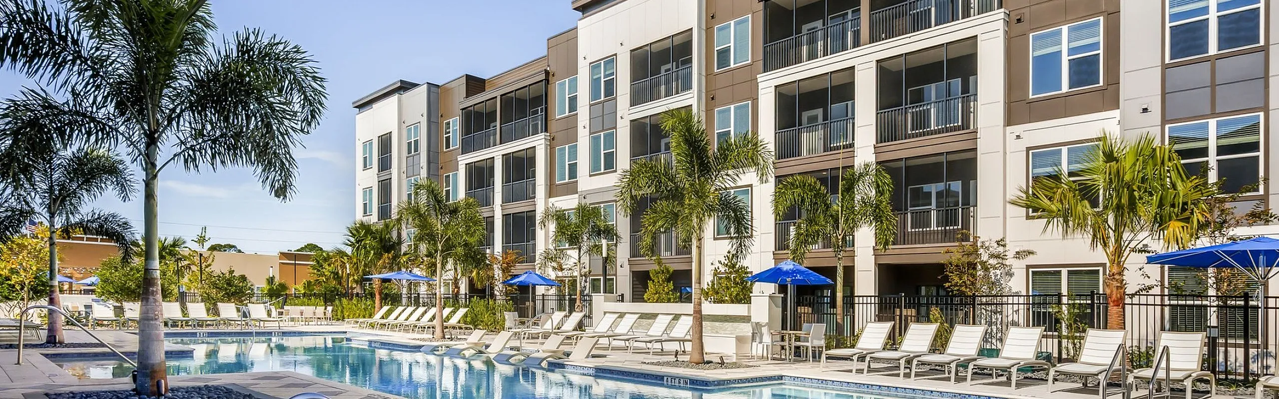 Luxurious apartment complex showcasing a sparkling blue swimming pool, surrounded by elegant sun loungers and tropical palm trees, set against modern architecture under a clear blue sky.