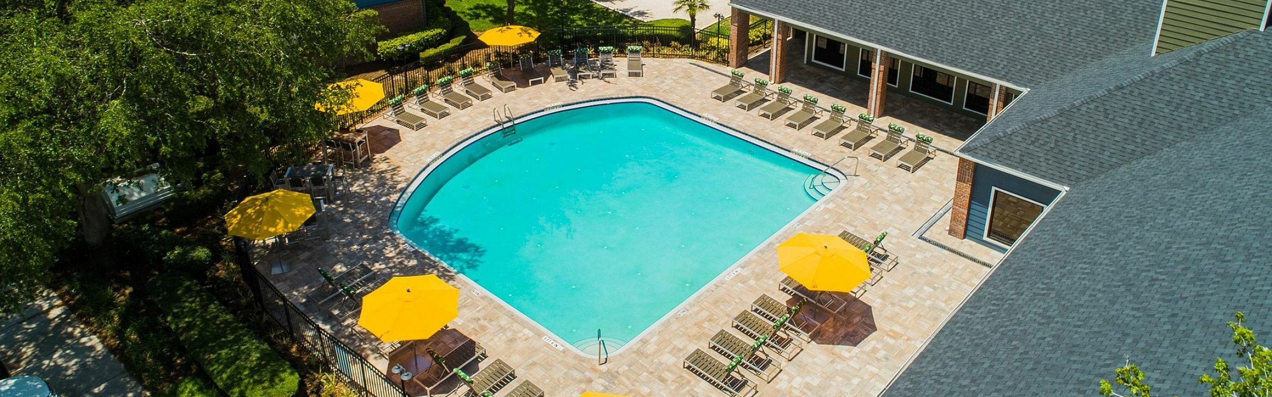 Vibrant aerial view of a resort-style swimming pool surrounded by lush greenery and charming buildings, featuring bright yellow umbrellas and ample sun loungers for relaxation.