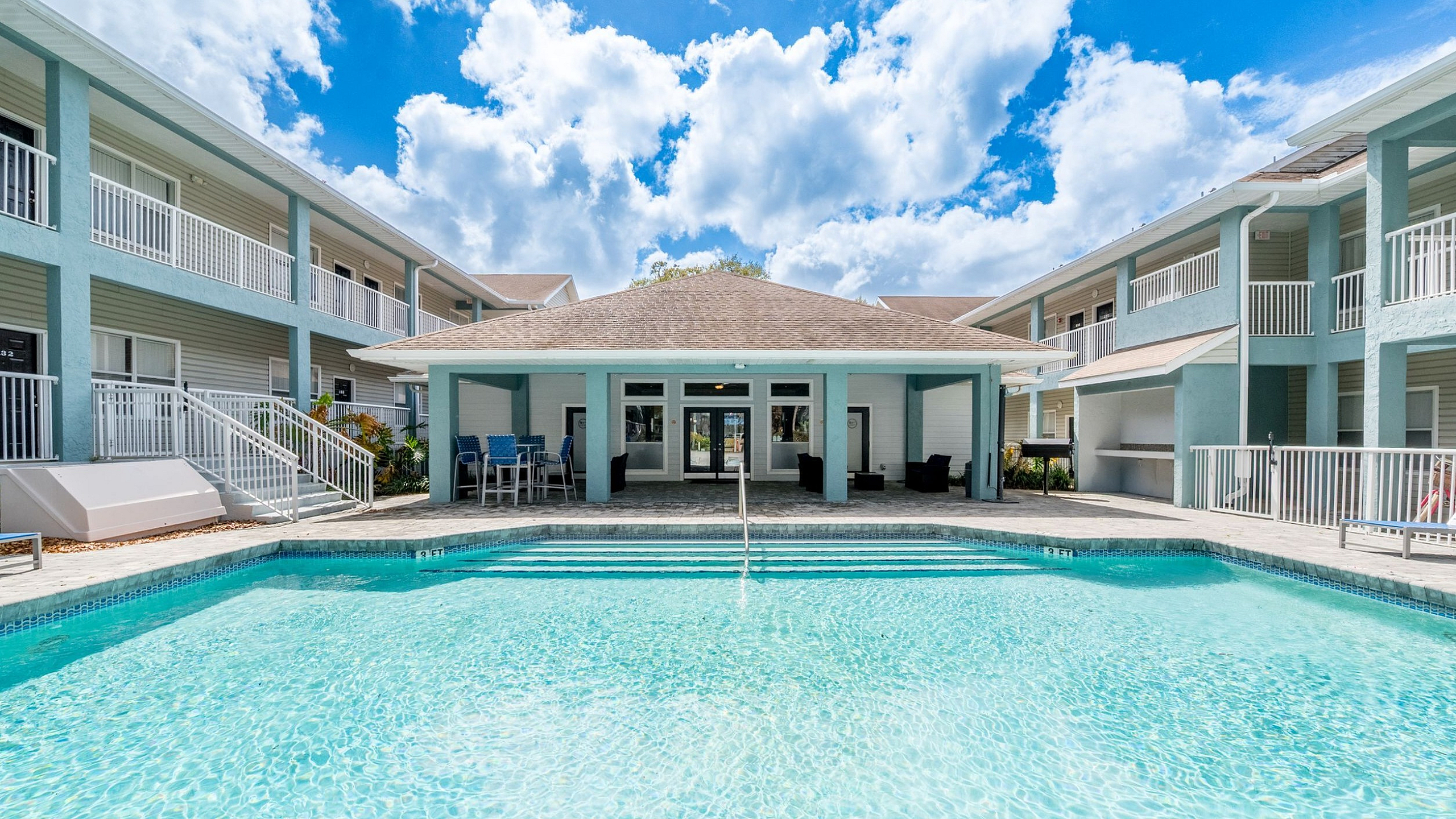 Sunny apartment complex featuring a sparkling blue pool, surrounded by modern two-story buildings with inviting balconies and a spacious patio area.