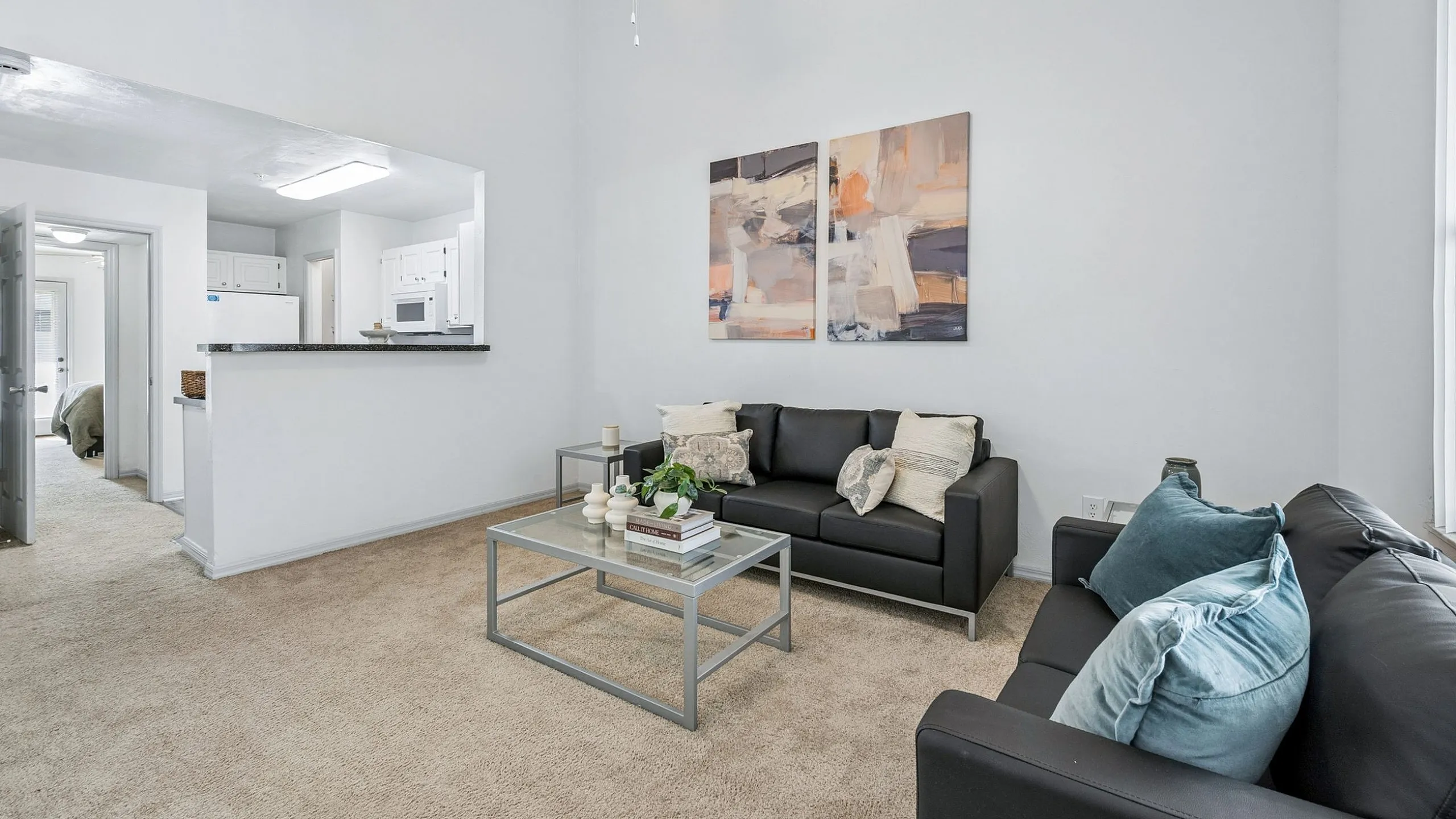 Bright and inviting open-concept living room with modern black sofas, abstract wall art, and a cozy carpet leading to a sleek kitchen area.
