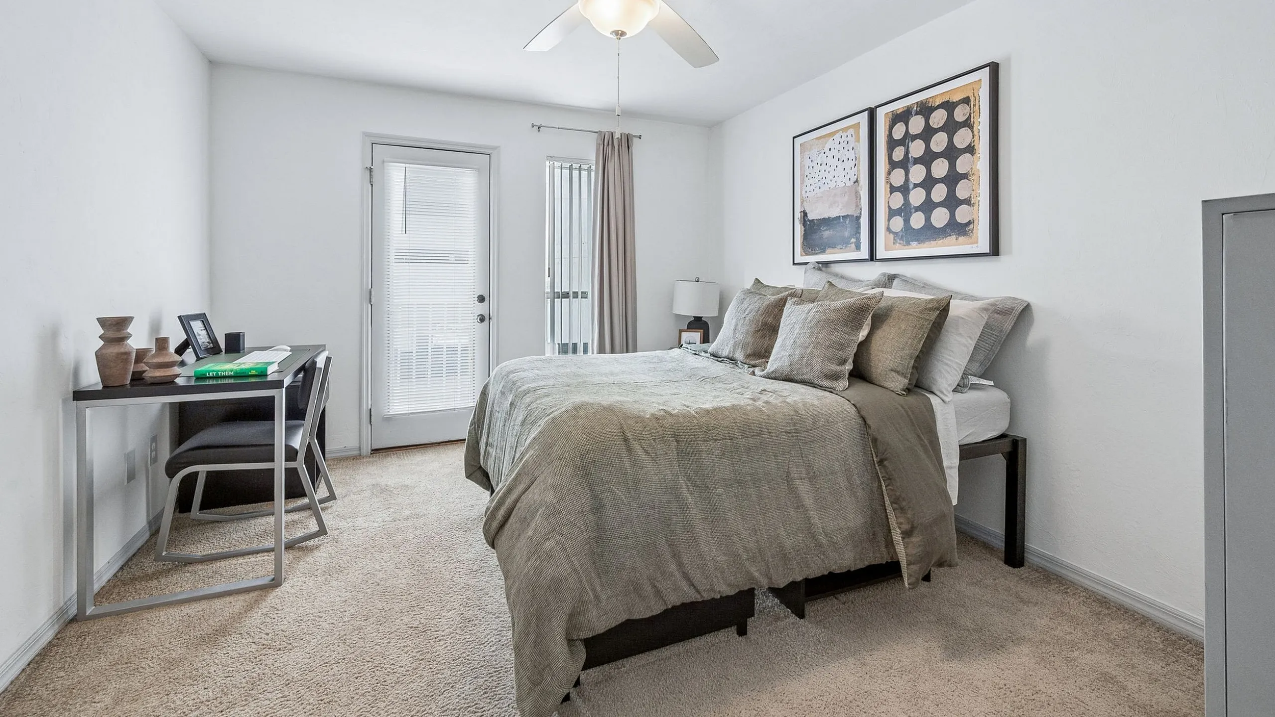 Bright and cozy bedroom featuring a comfortable bed, modern artwork, and a workspace with natural light streaming in through a glass door.