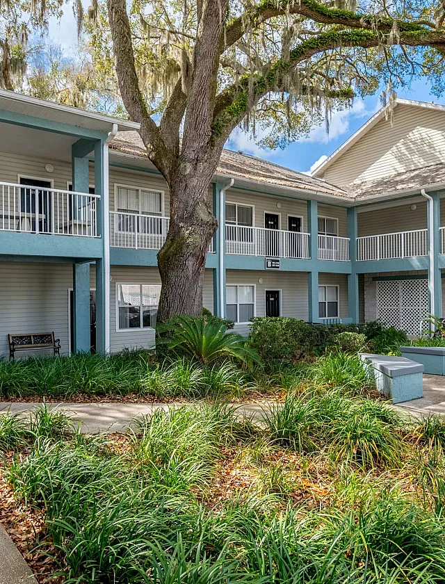 Charming apartment complex with vibrant landscaping, spacious balconies, and a welcoming courtyard under a lush canopy of trees.