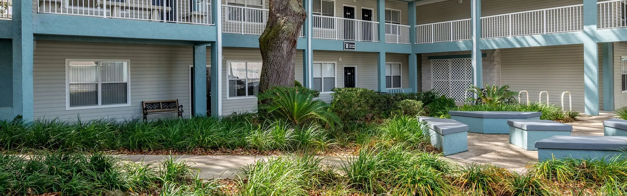 Charming two-story apartment complex with a spacious balcony, surrounded by lush greenery and outdoor seating, under a clear blue sky.