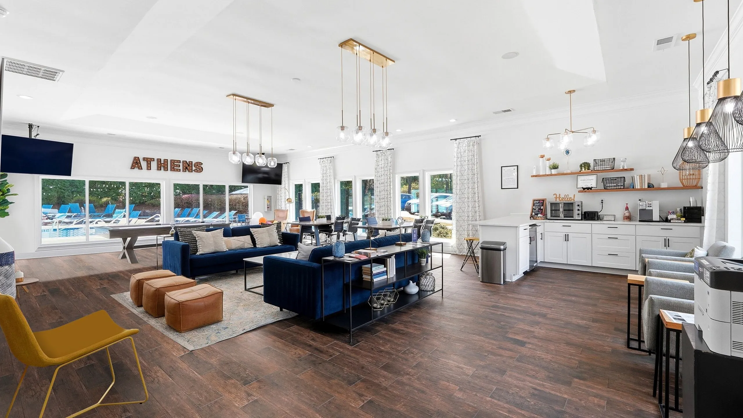 Chic communal lounge featuring elegant blue seating, stylish lighting fixtures, and a well-equipped kitchenette with a view of the outdoor pool area.