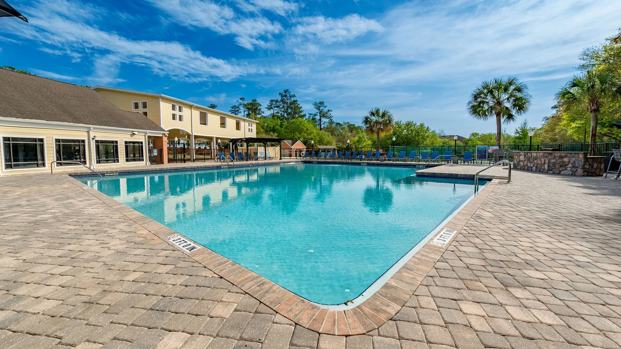 Inviting outdoor pool area with sparkling blue water, surrounded by scenic palm trees and a modern clubhouse under a clear sky.