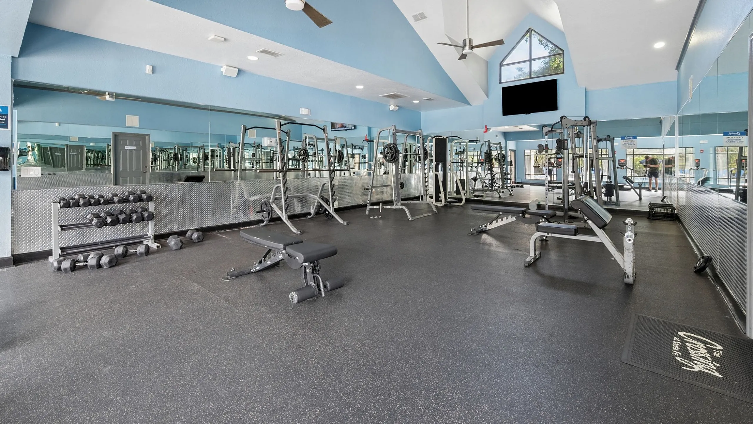 Bright and spacious gym with modern equipment, featuring large mirrors, blue accents, and ample natural light.