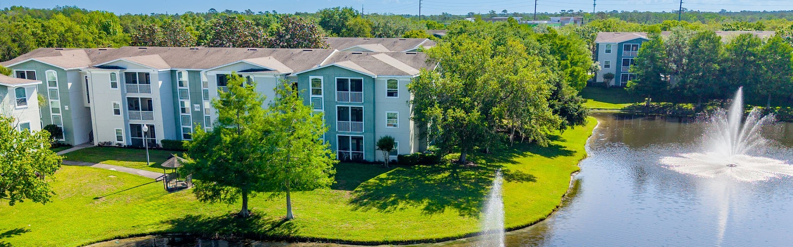 Scenic apartment complex with beautiful lake view and elegant water fountains surrounded by lush greenery.