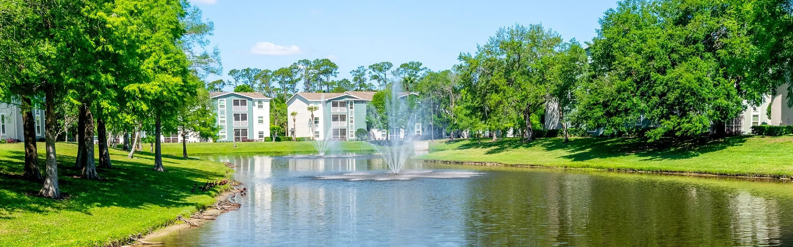 Serene lakeside view with lush greenery, elegant fountains, and modern apartments in the background, perfect for tranquil living.
