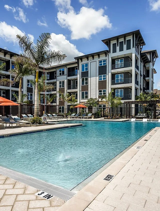 Luxurious apartment complex with modern architecture, highlighting a pristine swimming pool surrounded by palm trees and vibrant orange umbrellas under a clear blue sky.