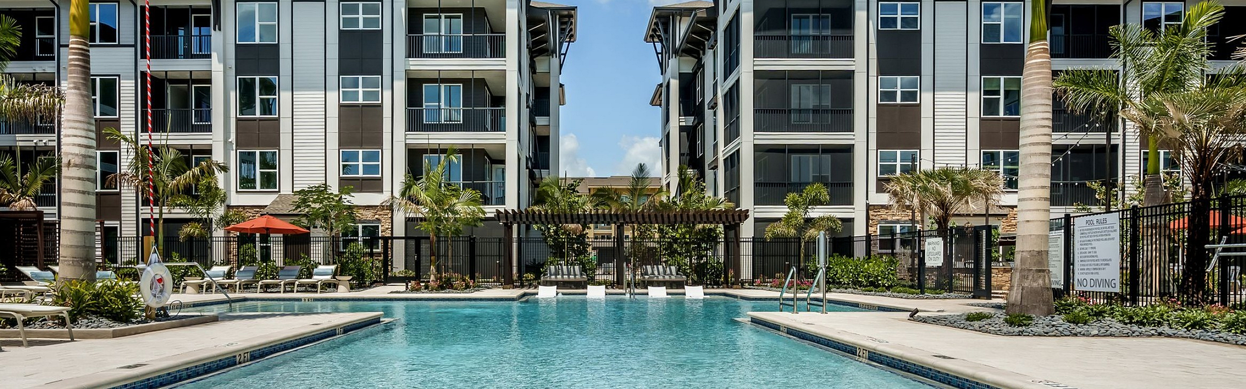 Luxurious apartment complex with a sparkling swimming pool, surrounded by lush palm trees and modern architecture for a resort-like living experience.