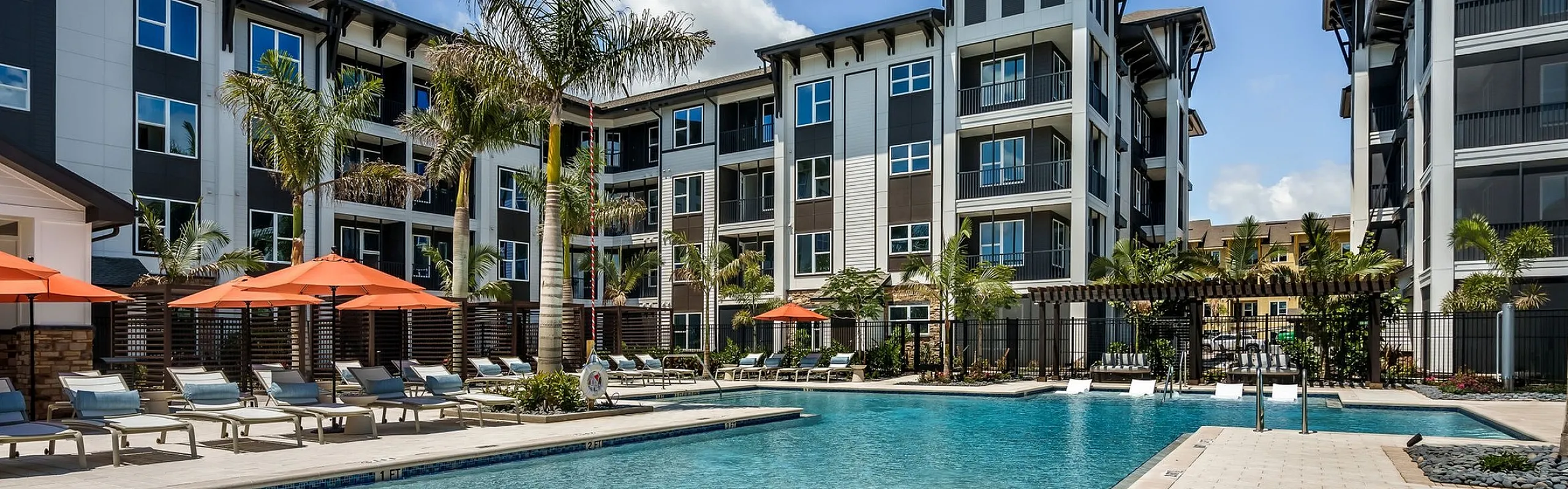 Luxurious apartment complex with a sparkling resort-style pool, surrounded by palm trees and stylish sun loungers under vibrant orange umbrellas.