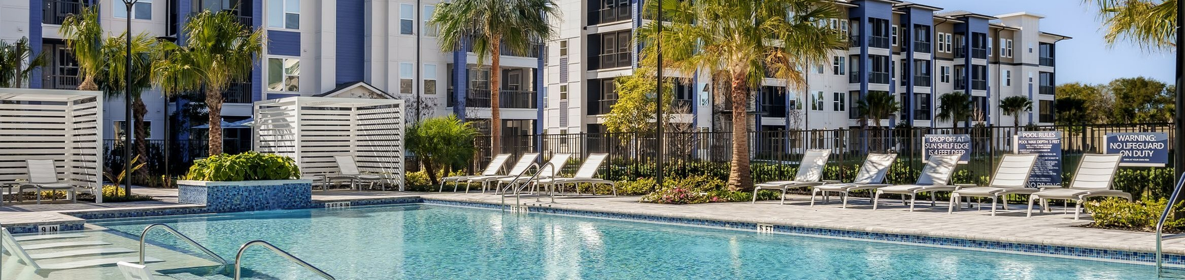 Luxurious apartment complex featuring a pristine swimming pool with lounge chairs and lush palm landscaping under a bright blue sky.