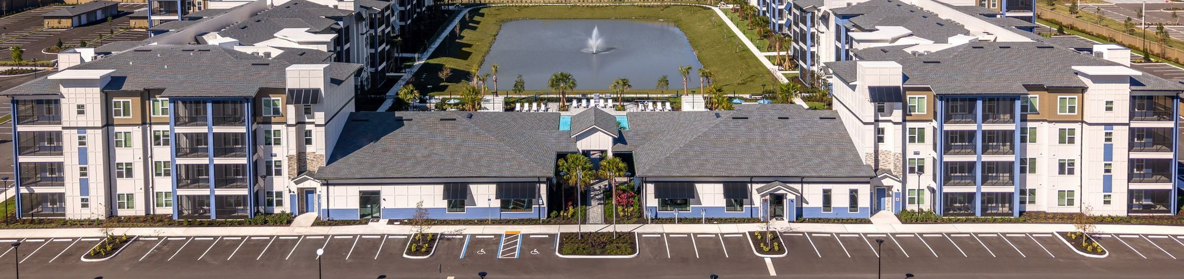 Aerial view of modern apartment complex with lush landscaping, central water feature, and ample parking, set against a picturesque backdrop.