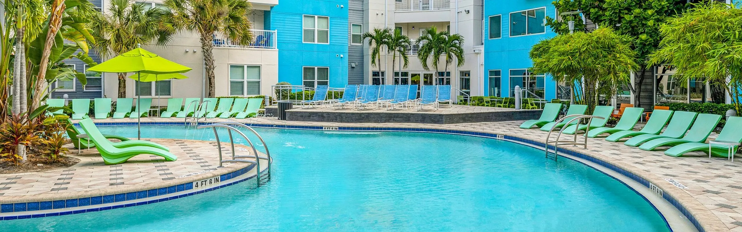 Vibrant apartment complex with a picturesque pool area, featuring lush tropical greenery and modern lounge chairs under bright blue skies.