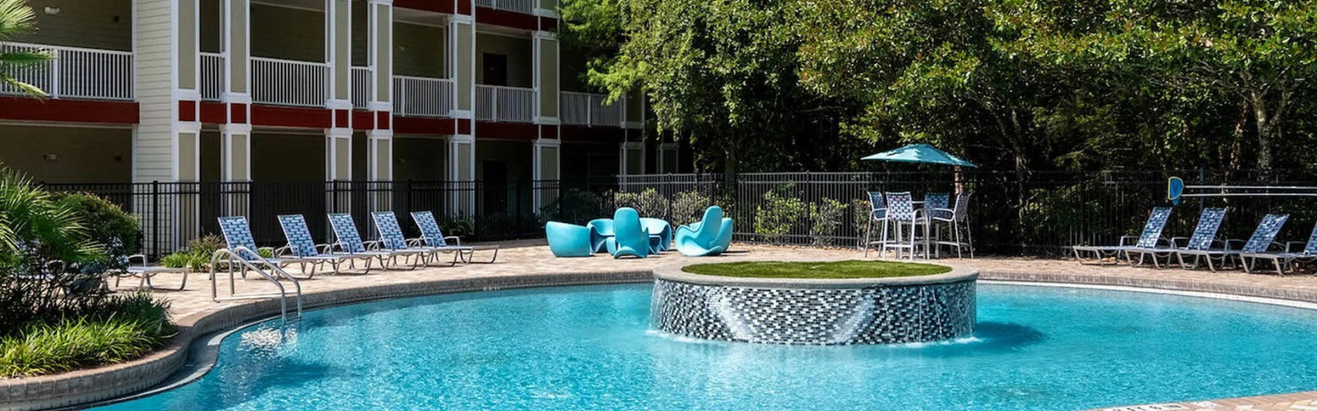 Inviting poolside view at a modern apartment complex, featuring a sparkling blue pool with lounge chairs and lush greenery.