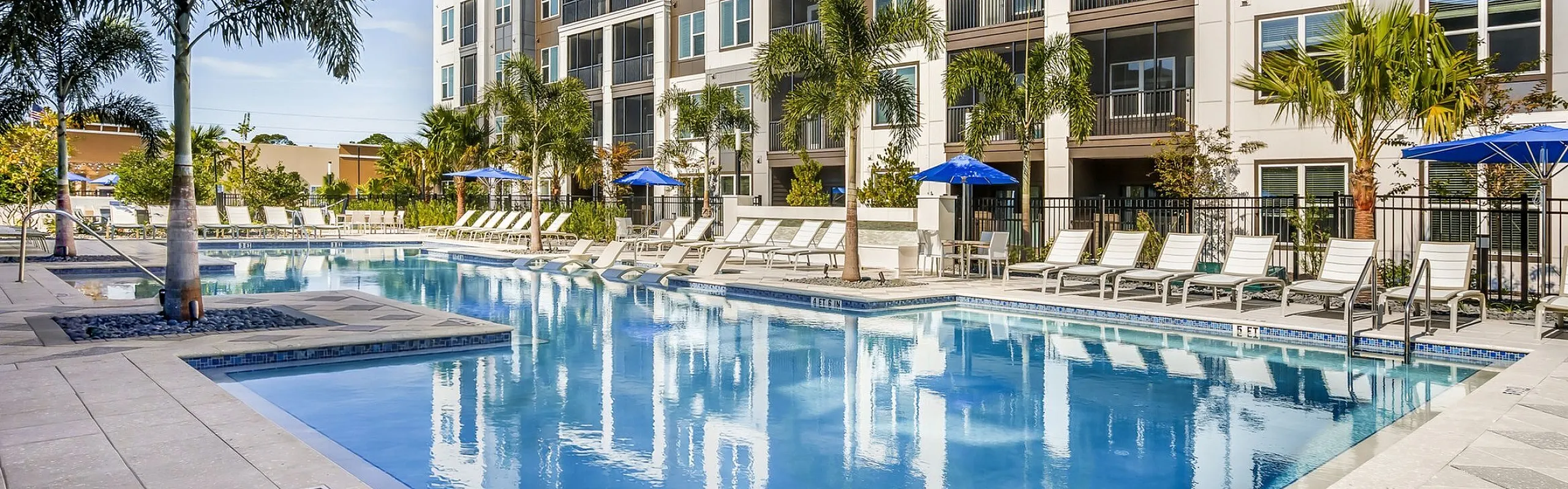 Luxurious apartment complex with modern exterior, featuring a large swimming pool, palm trees, and ample outdoor seating. Perfect for relaxation and leisure.