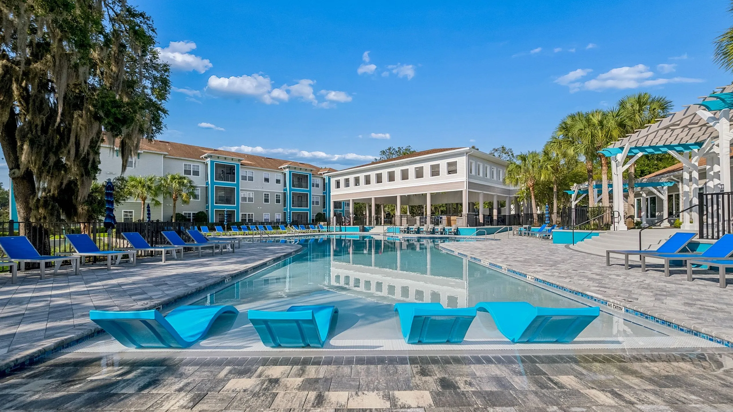 Luxurious apartment complex with a stunning outdoor pool, elegant lounge chairs, and lush palm landscaping under a bright blue sky.