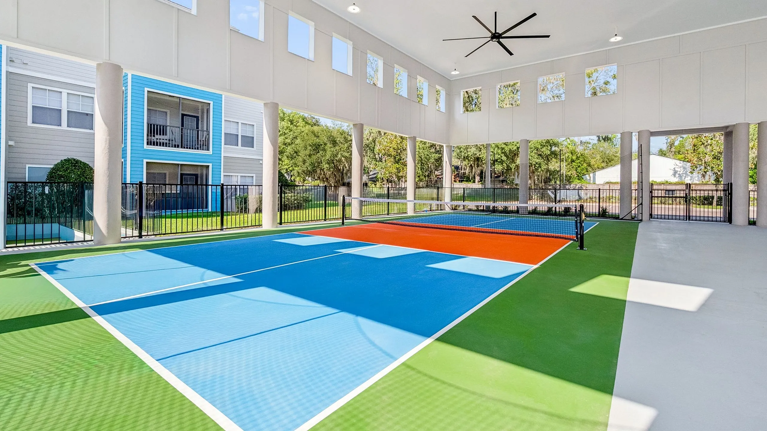 Modern indoor sports court with vibrant colors and large ceiling fan, surrounded by lush greenery and stylish apartments.