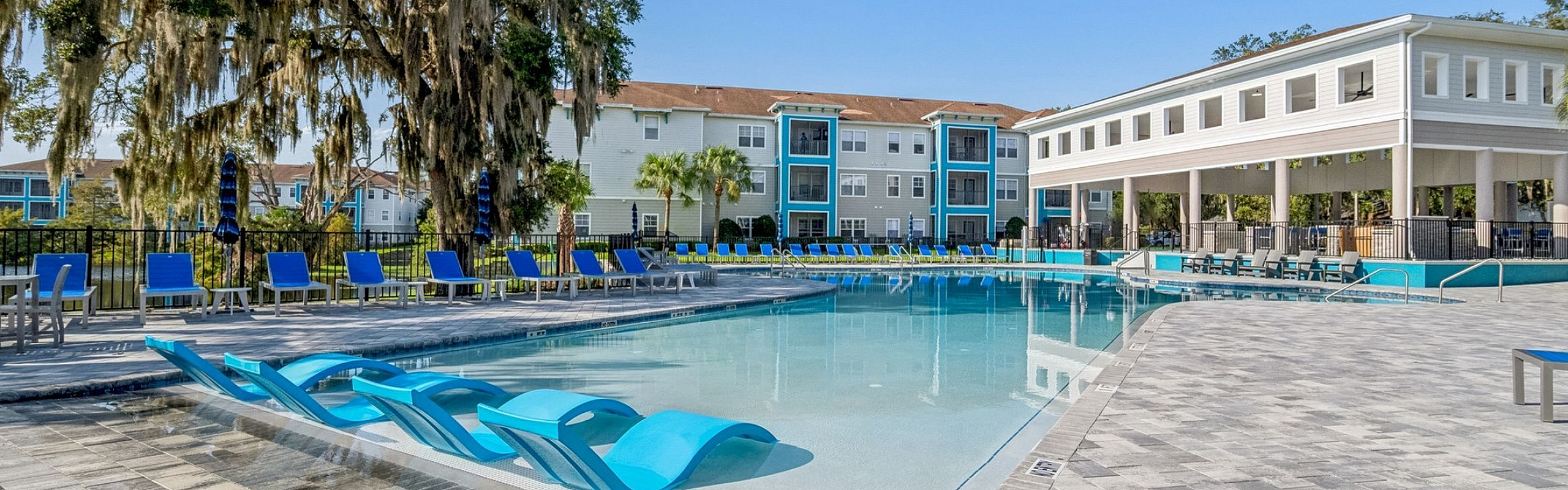 Chic apartment complex with expansive pool area, stylish lounging chairs, and lush greenery under a clear blue sky.