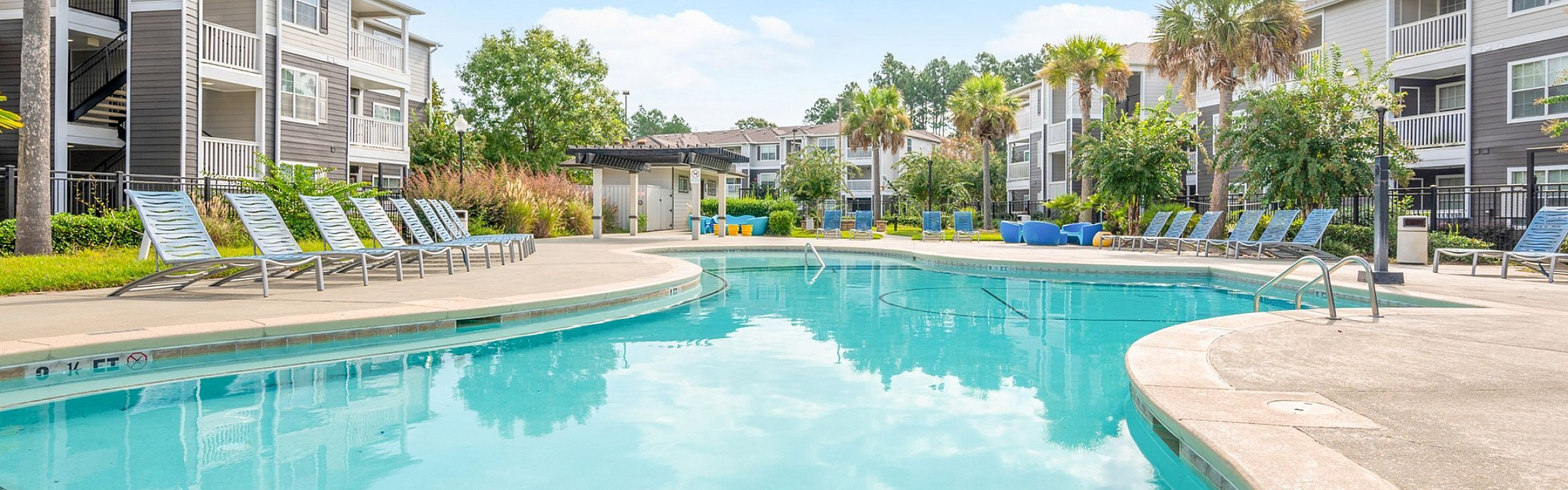 Luxurious apartment complex with a stunning outdoor pool and ample lounge seating, surrounded by lush greenery and modern architecture.