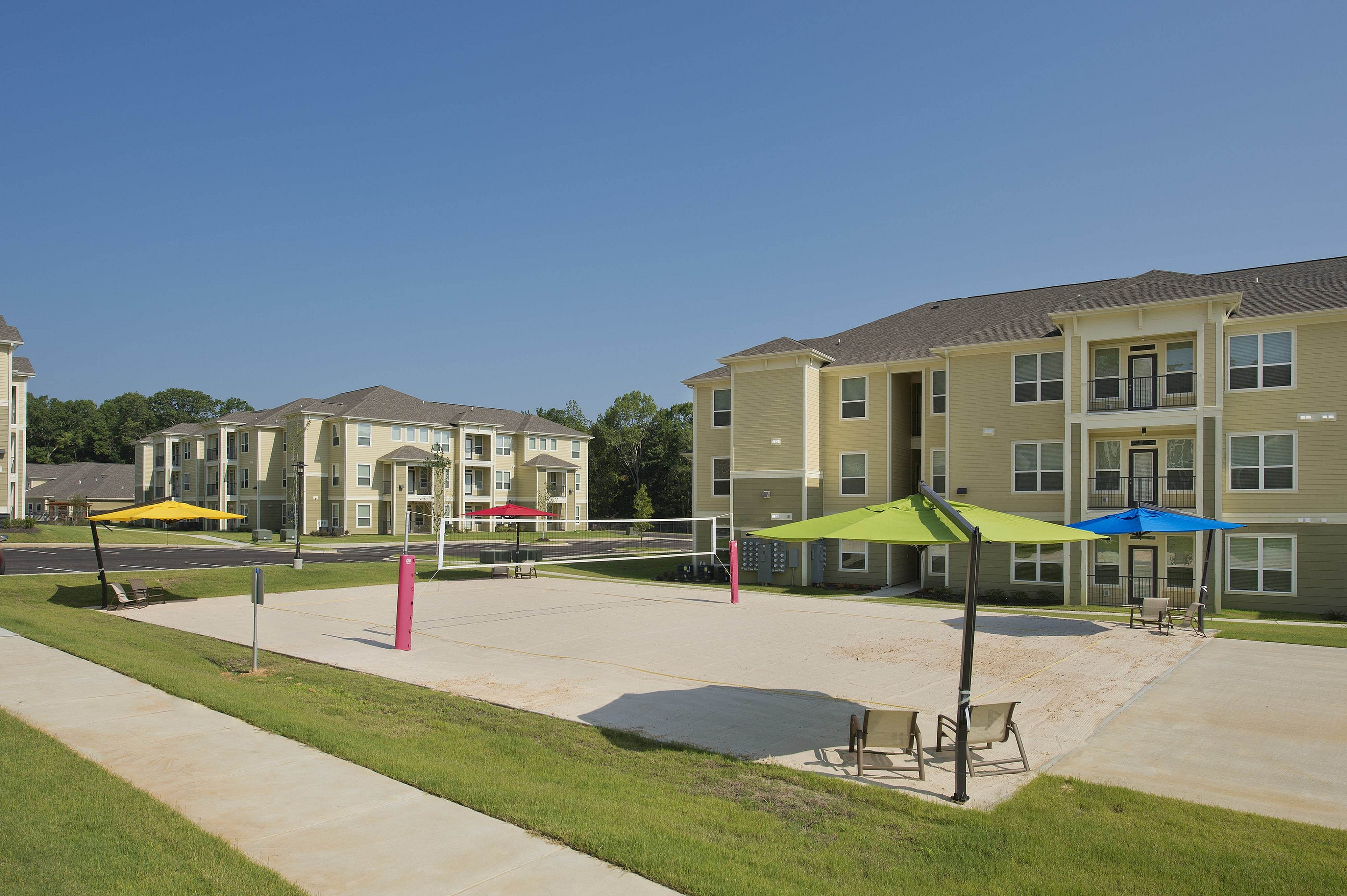 Charming apartment complex with a vibrant outdoor sand volleyball court and colorful parasols, surrounded by lush greenery and modern architecture.