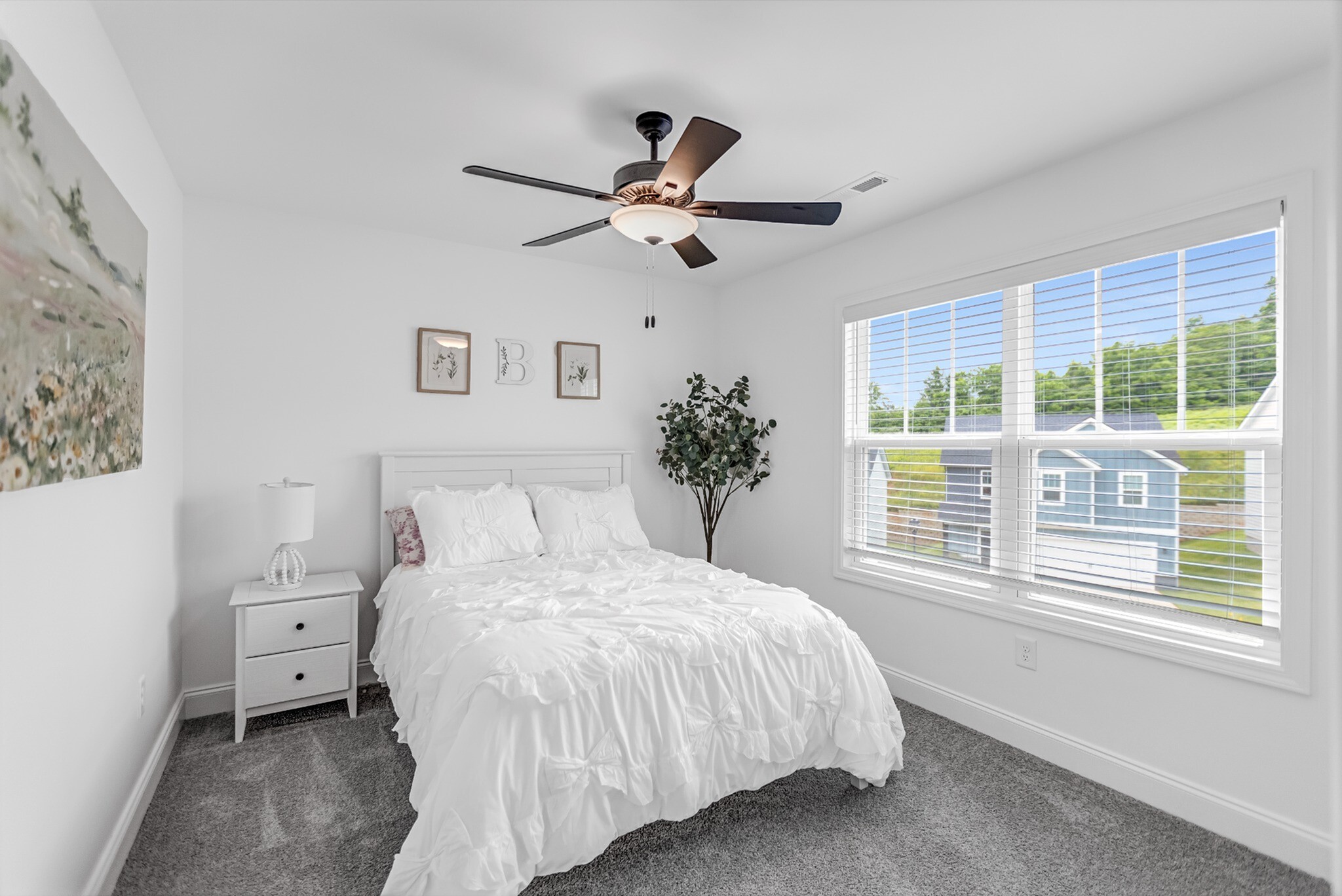 Serene bedroom with plush white bedding, modern ceiling fan, and large window offering bright natural lighting and charming exterior views.