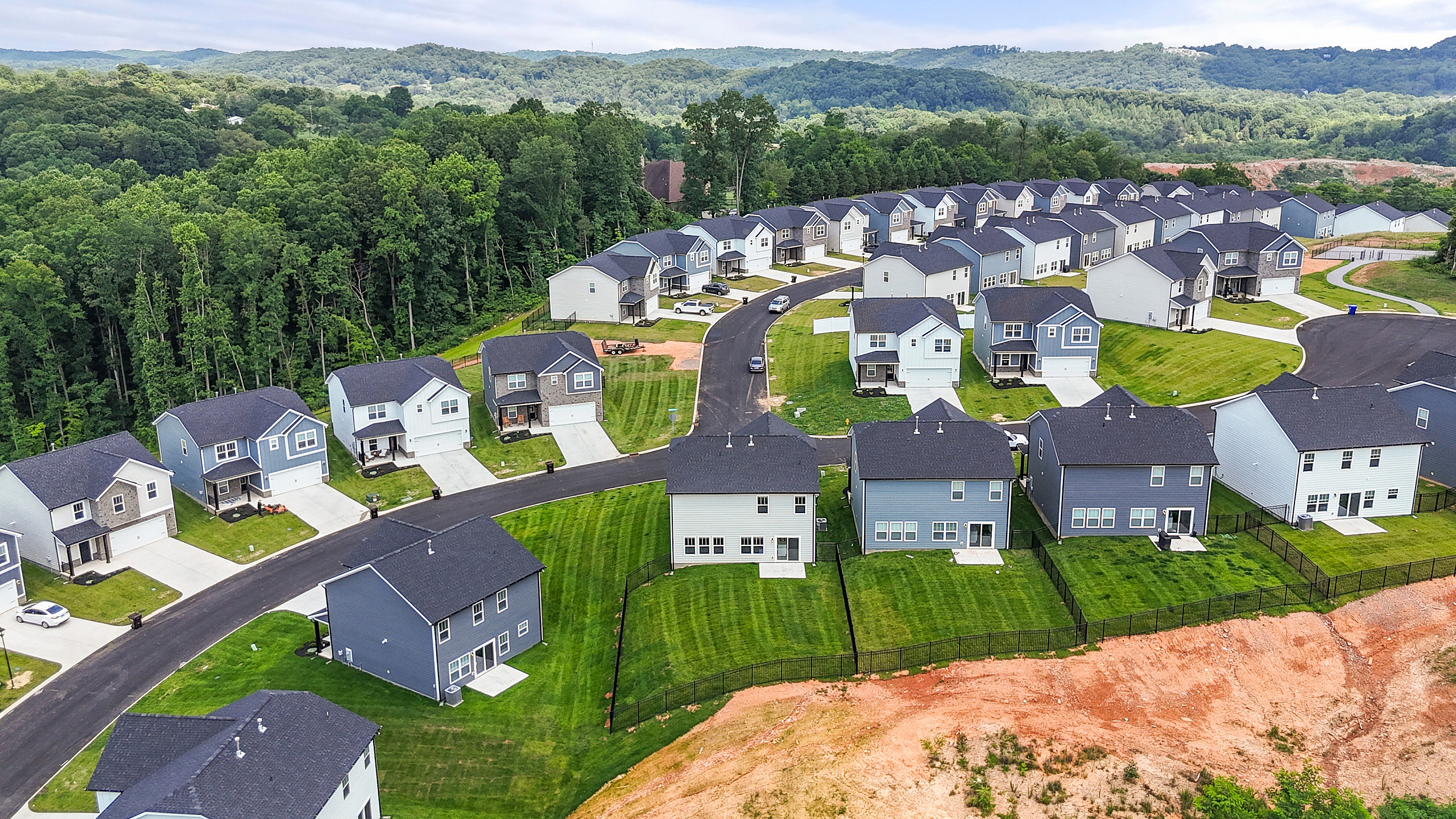 Aerial view of a picturesque suburban neighborhood surrounded by lush forests, featuring charming two-story homes with manicured lawns and scenic mountain backdrop.