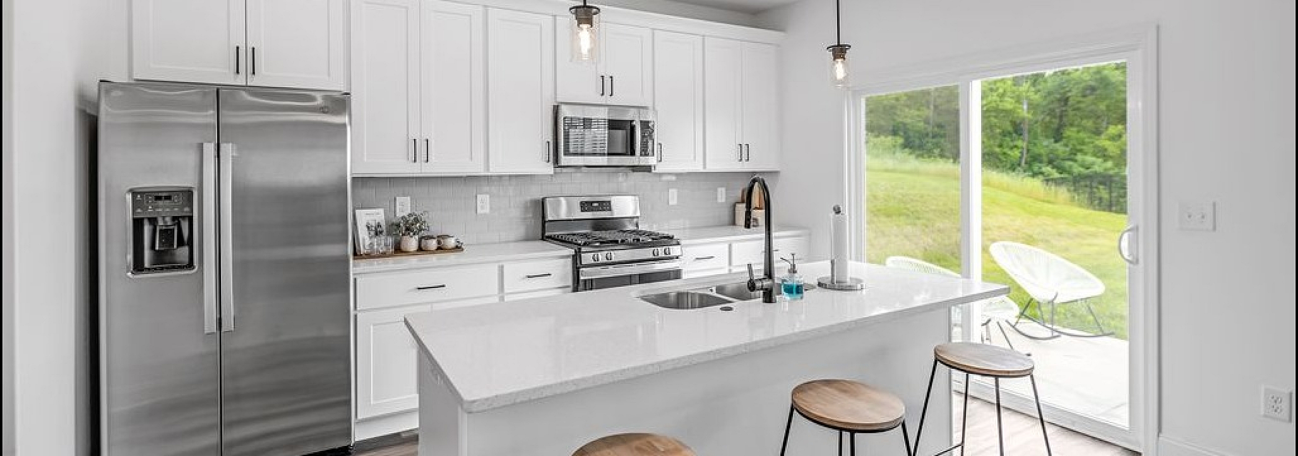 Modern kitchen with sleek white cabinetry, stainless steel appliances, and a central island with bar stools overlooking a lush green outdoor space.